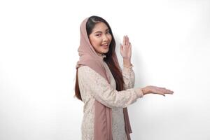 ásia muçulmano mulher vestindo lenço de cabeça véu hijab sorridente alegremente enquanto olhando e apontando para cópia de espaço ao lado dela, em pé em isolado branco fundo foto