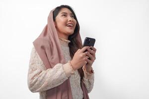 alegre jovem ásia muçulmano mulher dentro véu hijab sorridente e sentindo-me feliz enquanto segurando uma Smartphone a partir de lado visualizar. Ramadã e eid Mubarak conceito foto