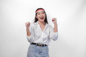 retrato do a animado jovem ásia mulher levantando punho acima gesto, ganhando e a comemorar vitória pose, vestindo bandeira arco de cabelo e branco camisa. independência dia conceito foto