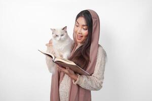 atraente jovem ásia muçulmano mulher dentro véu hijab sorridente enquanto abraçando uma branco boneca de pano gato animal e segurando al Alcorão em a de outros mão foto