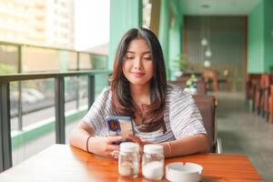 jovem ásia mulher vestindo vestir sentado às uma restaurante para uma café da manhã, olhando às Smartphone dentro uma profundo pensamento com sério face foto