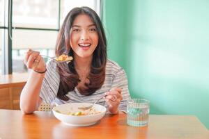 retrato do a animado jovem ásia mulher vestindo vestir sentado às uma restaurante, comendo e desfrutando café da manhã com uma alegre sorrir foto