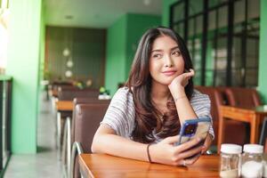 pensativo jovem ásia mulher vestindo vestir sentado às uma restaurante para uma café da manhã, segurando uma Smartphone enquanto escoramento dela queixo foto