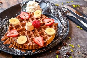 morango e banana waffle com chocolate, açoitado creme, pistache, faca e garfo servido dentro prato isolado em Sombrio fundo topo Visão cafeteria sobremesa Comida foto