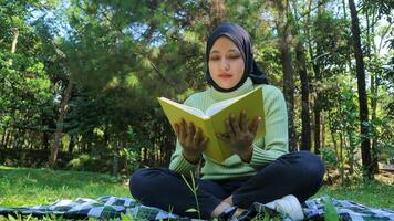 relaxado muçulmano mulher desfrutando final de semana às parque, sentado em Relva e lendo livro, esvaziar espaço foto