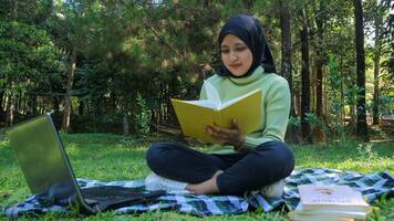 relaxado muçulmano mulher desfrutando final de semana às parque, sentado em Relva e lendo livro, esvaziar espaço foto