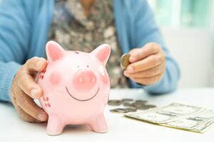 ásia idosos mulher colocando moeda para dentro Rosa porquinho banco para salvando dinheiro e seguro, pobreza, financeiro problema dentro aposentadoria. foto