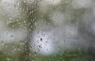 uma foto de chuva cai no vidro da janela com uma visão turva das árvores verdes florescendo. imagem abstrata mostrando condições de tempo nublado e chuvoso