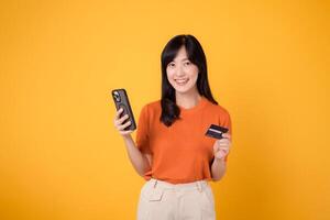animado jovem ásia mulher 30s dentro laranja camisa, usando Smartphone e segurando crédito cartão em amarelo fundo. agradável conectados compras. foto