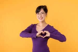 retrato alegre jovem ásia 30s mulher feliz sorrir vestindo roxa suéter camisa expressando coração mão placa gesto em peito isolado amarelo fundo. namorados romântico símbolo publicidade conceito. foto