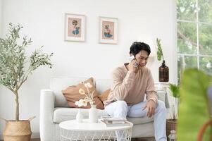 feliz ásia homem sentado em sofá usando Smartphone dentro vivo quarto às lar, relaxar Tempo e estilo de vida conceito foto