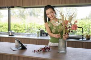 ásia sorridente mulher colocando lindo flores em jarra , desfrutando decoração às casa foto