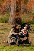 jovem mulher com fofa bebê menina dentro bebê carrinho de criança às a outono parque foto