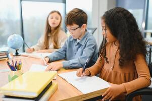 focado multirracial alunos crianças escrevendo baixa dados para dentro caderno enquanto sentado às mesa foto