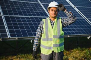 retrato do jovem indiano homem técnico vestindo branco Difícil chapéu em pé perto solar painéis contra azul céu. industrial trabalhador solar sistema instalação foto