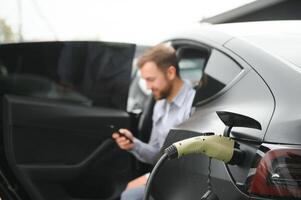 feliz homem usando inteligente telefone e cobrando carro às veículo cobrando estação foto