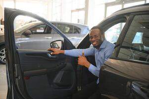 carro proprietário. alegre afro cara sorridente, sentado dentro Novo automóvel dirigindo a partir de concessionária fazer compras foto