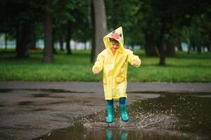 pequeno Garoto jogando dentro chuvoso verão parque. criança com guarda-chuva, à prova d'água casaco e chuteiras pulando dentro poça e lama dentro a chuva. criança caminhando dentro verão chuva ao ar livre Diversão de qualquer clima. feliz infância. foto