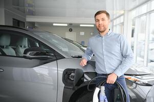 bonito o negócio homem segurando cobrando cabo para elétrico carro. caucasiano masculino carrinhos perto elétrico auto dentro concessionária foto