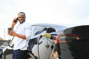 africano homem em pé perto elétrico carro com cobrando cabo dentro plugue. eco amigáveis veículo cobrando em estação. estilos de vida conceito foto