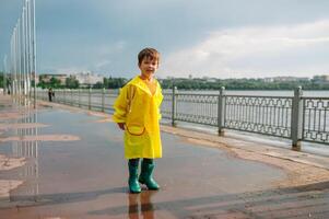 pequeno Garoto jogando dentro chuvoso verão parque. criança com guarda-chuva, à prova d'água casaco e chuteiras pulando dentro poça e lama dentro a chuva. criança caminhando dentro verão chuva ao ar livre Diversão de qualquer clima. feliz infância. foto