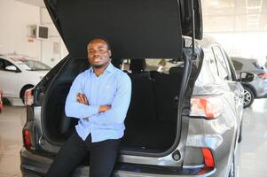 feliz carro comprador, Novo carro proprietário conceito. retrato do animado jovem africano americano cara dentro concessionária sala de exposições foto