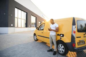 casual africano homem com Smartphone perto elétrico carro esperando para a terminar do a bateria cobrando processo. foto