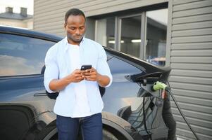 feliz jovem homem com Móvel telefone cobrando carro às elétrico veículo cobrando estação foto