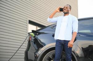 a africano homem é em pé perto a elétrico carro, esperando para isto para carregar às uma cobrando estação e usando a telefone. foto