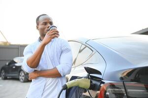 africano homem em pé perto elétrico carro com cobrando cabo dentro plugue. eco amigáveis veículo cobrando em estação. estilos de vida conceito foto