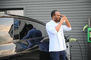 africano homem em pé perto elétrico carro com cobrando cabo dentro plugue. eco amigáveis veículo cobrando em estação. estilos de vida conceito foto