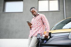 casual africano homem com Smartphone perto elétrico carro esperando para a terminar do a bateria cobrando processo. foto