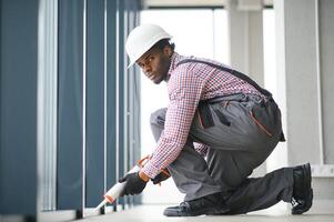 baixo ângulo Visão do uma jovem africano reparador dentro macacão instalando janela foto