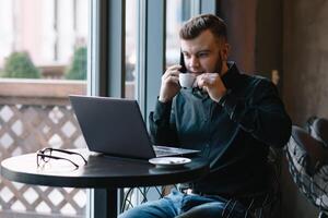 jovem homem de negocios falando em Móvel telefone enquanto trabalhando em computador portátil dentro cafeteria foto