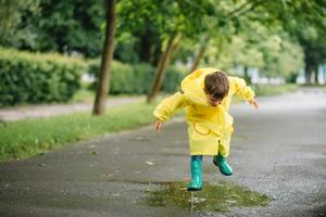 pequeno Garoto jogando dentro chuvoso verão parque. criança com guarda-chuva, à prova d'água casaco e chuteiras pulando dentro poça e lama dentro a chuva. criança caminhando dentro verão chuva ao ar livre Diversão de qualquer clima. feliz infância foto