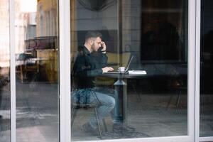 jovem homem de negocios falando em Móvel telefone enquanto trabalhando em computador portátil dentro cafeteria. foto