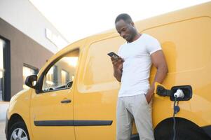 casual africano homem com Smartphone perto elétrico carro esperando para a terminar do a bateria cobrando processo. foto