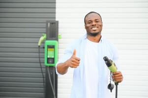 sério africano homem segurando carregar cabo dentro em mão, em pé perto luxo elétrico carro. foto