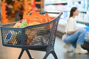 uma cheio cesta do produtos dentro uma supermercado em a fundo do uma jovem mulher foto