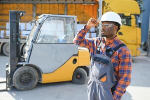 retrato do uma feliz africano americano masculino trabalhador dirigindo empilhadeira às local de trabalho foto