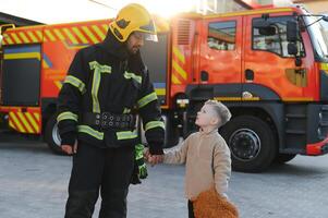 uma bombeiro levar uma pequeno criança Garoto para Salve  ele. fogo motor carro em fundo foto