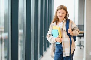 pequeno lindo escola menina em pé entre corredor às escola, segurando notas às mãos. engraçado e feliz menina sorridente às Câmera, em repouso depois de lições em primário escola foto