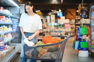 jovem satisfeito Diversão mulher dentro casual roupas compras às supermercado loja com mercearia carrinho foto