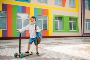 Adolescência Garoto com pontapé lambreta perto moderno escola. criança com mochila e livro ao ar livre. começando do aulas. primeiro dia do cair. costas para escola. foto