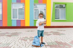 costas para escola. feliz sorridente Garoto dentro óculos é indo para escola para a primeiro tempo. criança com mochila e livro ao ar livre. começando do aulas. primeiro dia do cair. foto