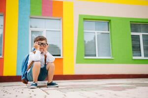 triste pequeno Garoto lado de fora do escola. triste estudante com livros perto uma moderno escola. escola conceito. costas para escola foto