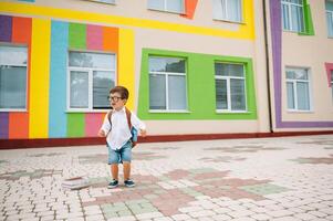 costas para escola. feliz sorridente Garoto dentro óculos é indo para escola para a primeiro tempo. criança com mochila e livro ao ar livre. começando do aulas. primeiro dia do outono foto