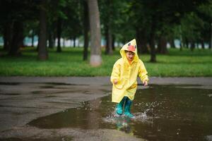 pequeno Garoto jogando dentro chuvoso verão parque. criança com guarda-chuva, à prova d'água casaco e chuteiras pulando dentro poça e lama dentro a chuva. criança caminhando dentro verão chuva ao ar livre Diversão de qualquer clima. feliz infância. foto