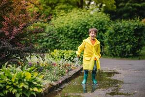 pequeno Garoto jogando dentro chuvoso verão parque. criança com guarda-chuva, à prova d'água casaco e chuteiras pulando dentro poça e lama dentro a chuva. criança caminhando dentro verão chuva ao ar livre Diversão de qualquer clima. feliz infância. foto