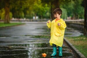 criança jogando com brinquedo barco dentro poça. criança jogar ao ar livre de chuva. outono chuvoso clima ao ar livre atividade para jovem crianças. criança pulando dentro turvar poças. à prova d'água Jaqueta e chuteiras para bebê. infância foto